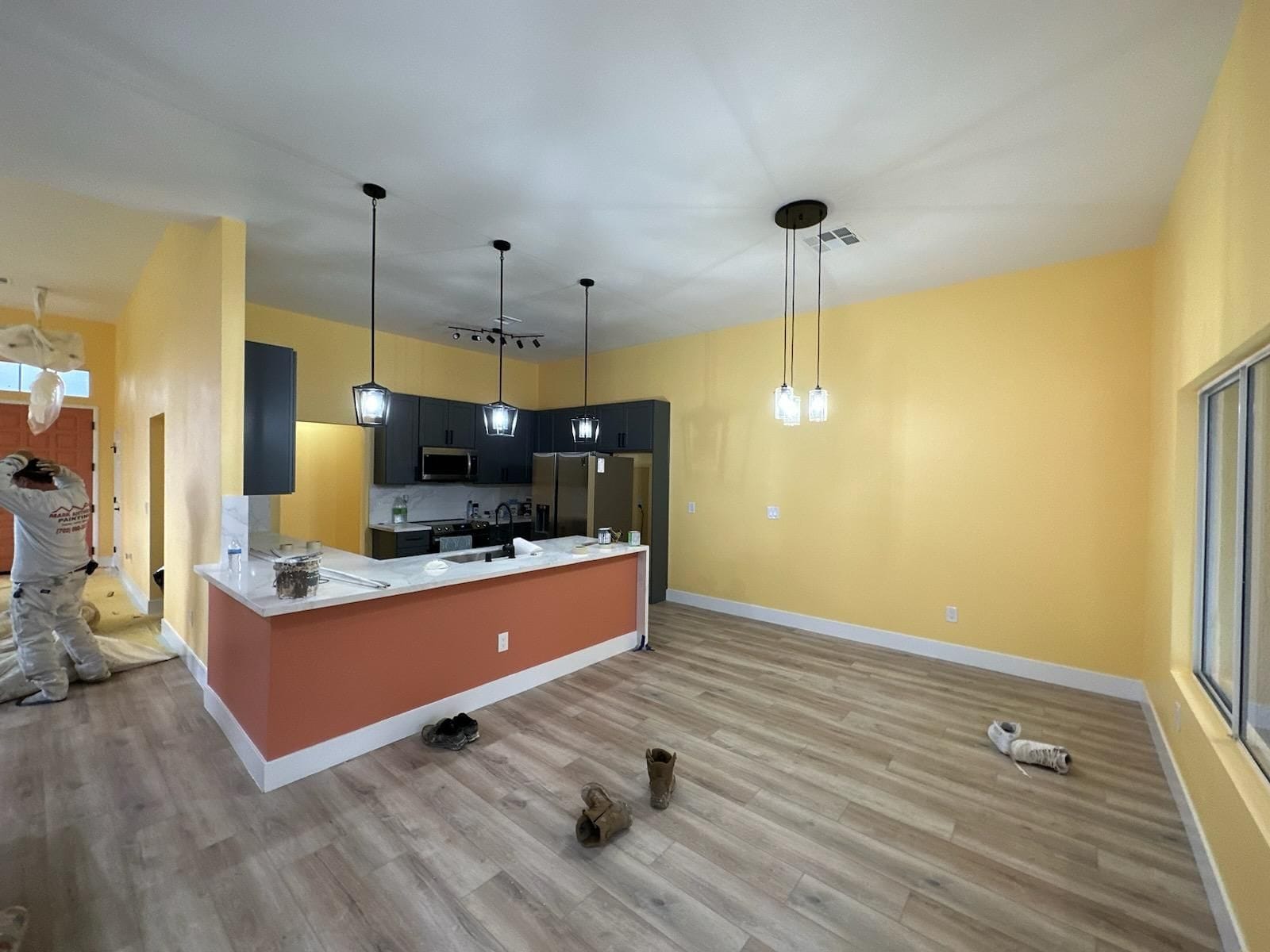 Open-concept kitchen with yellow walls, orange island, wood floors, and pendant lights. A person is painting in the doorway.