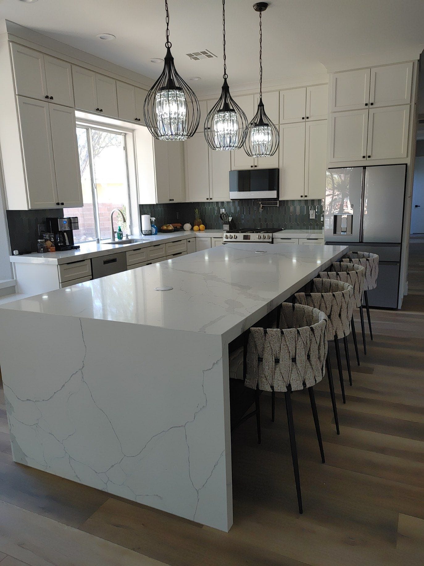 Modern kitchen with large white island, pendant lights, and bar stools. Light wood floors and stainless steel appliances.