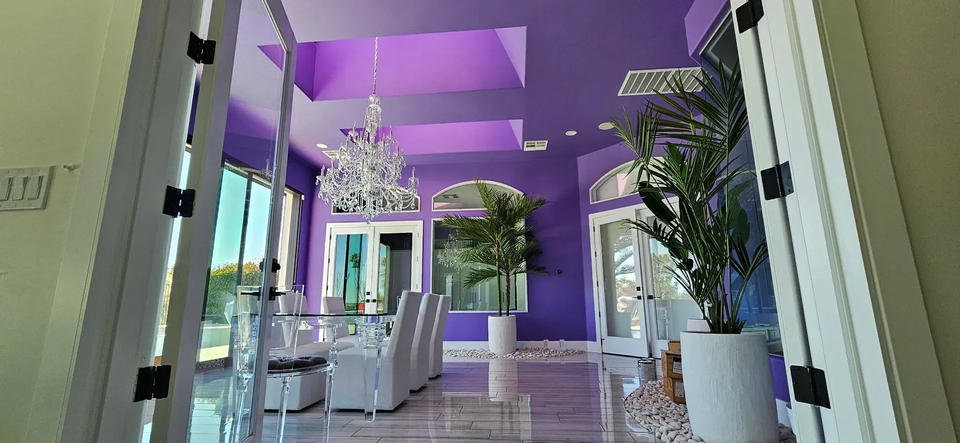 Purple dining room with a chandelier, white furniture, and large plants.