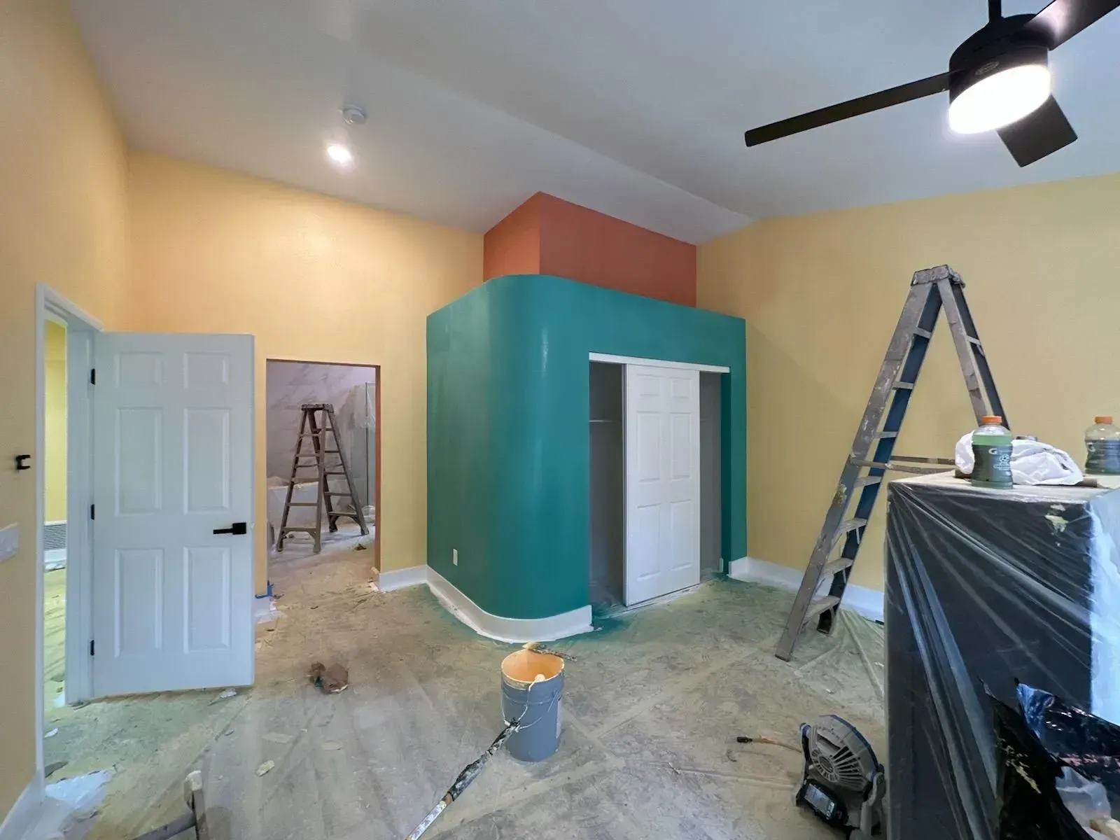 Room interior during renovation. Walls are painted yellow, teal, and coral. Ladder and tools are present.