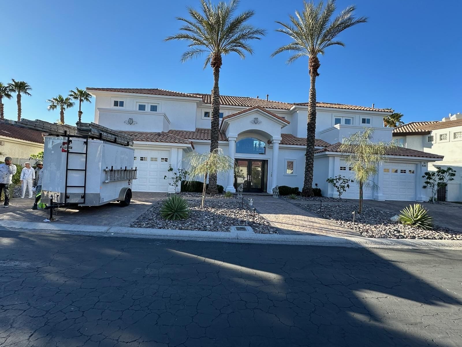White stucco house with three-car garage and palm trees, a trailer in the driveway, blue sky.
