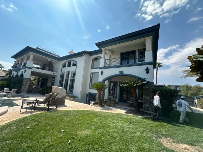 Large white house with green trim, lawn, pool area, and two people working on the grass under a bright sunny sky.