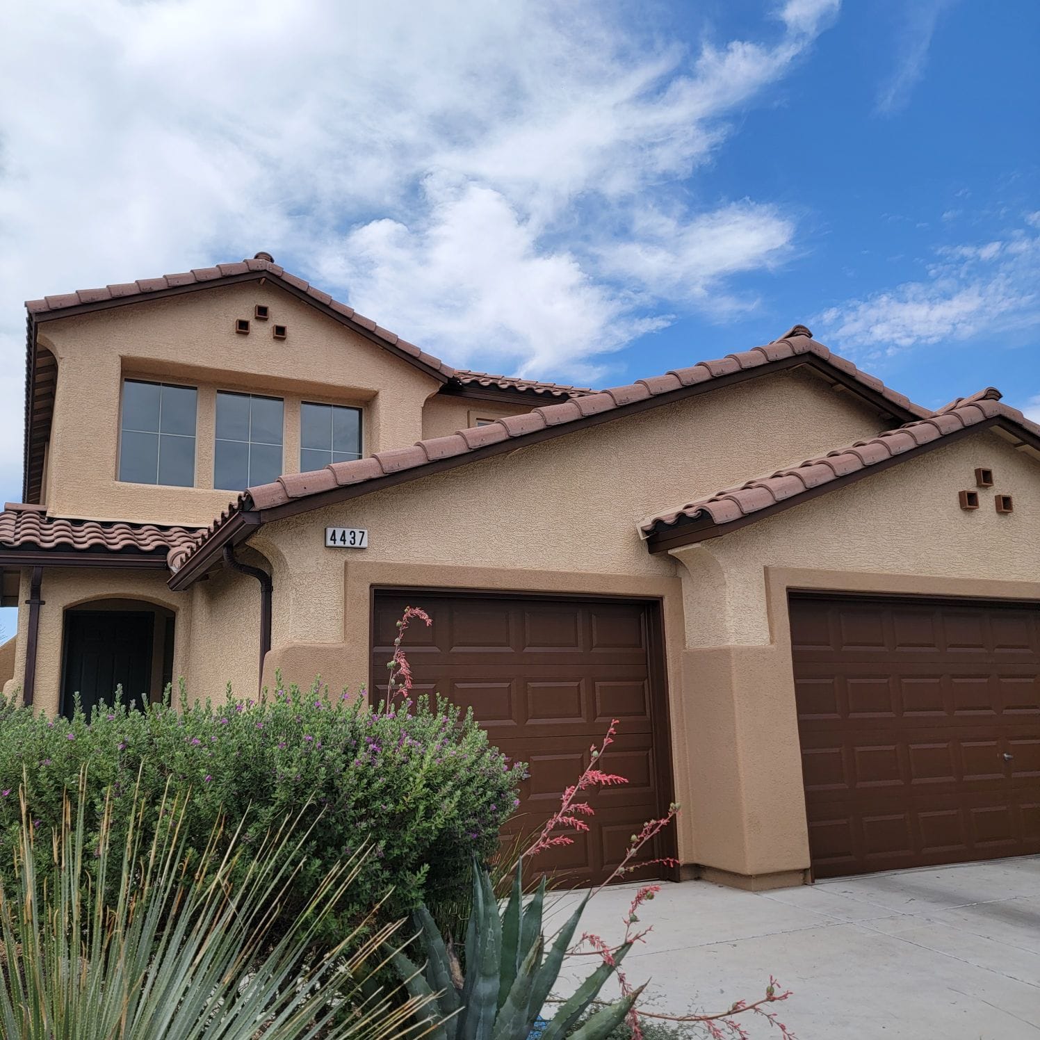 Tan two-story house with brown roof and garage doors against a blue sky.