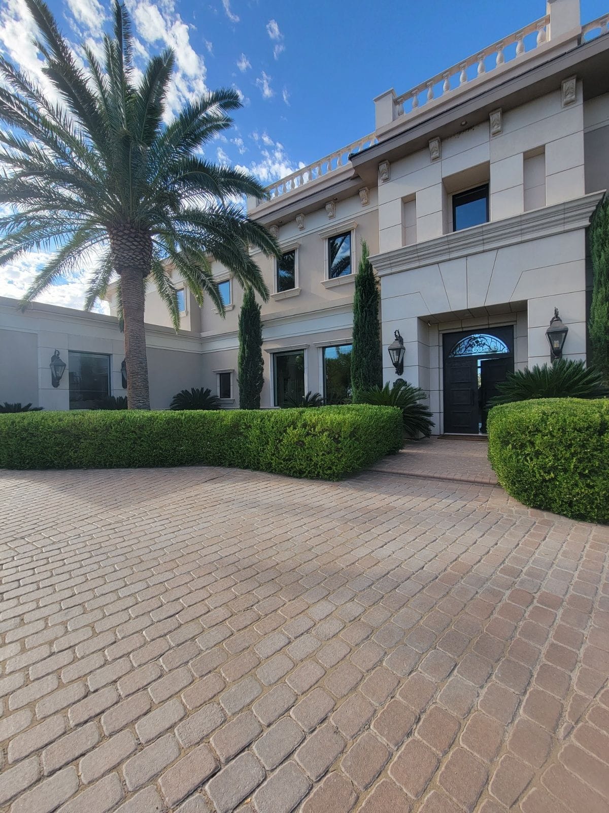 Large beige mansion with brick driveway, palm tree, and manicured hedges under a blue sky.