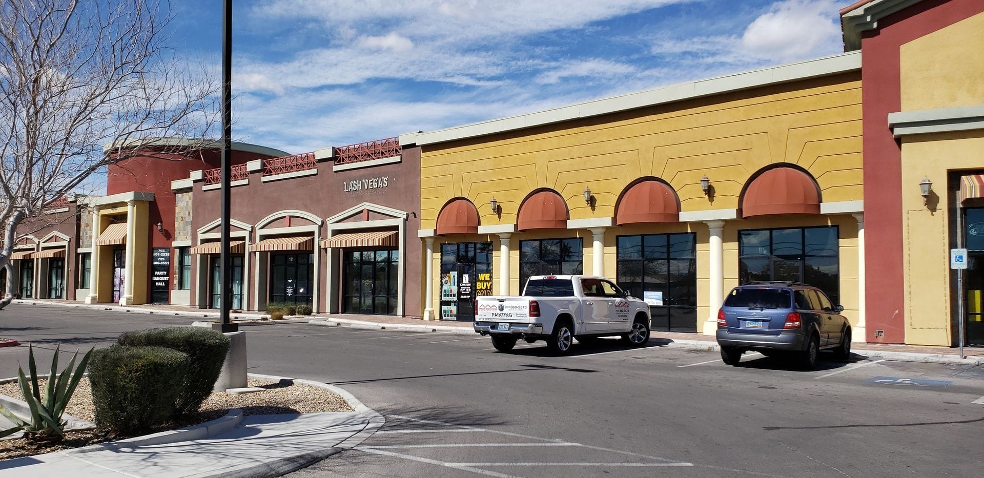 Shopping center with several stores. White truck and blue car are parked in front.